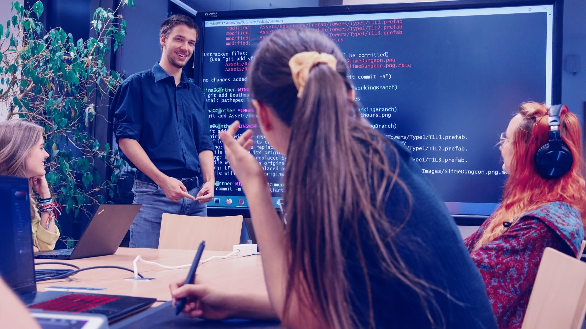 Personen im Gespräch vor einem großen Bildschirm auf dem Programmiercode zus ehen ist