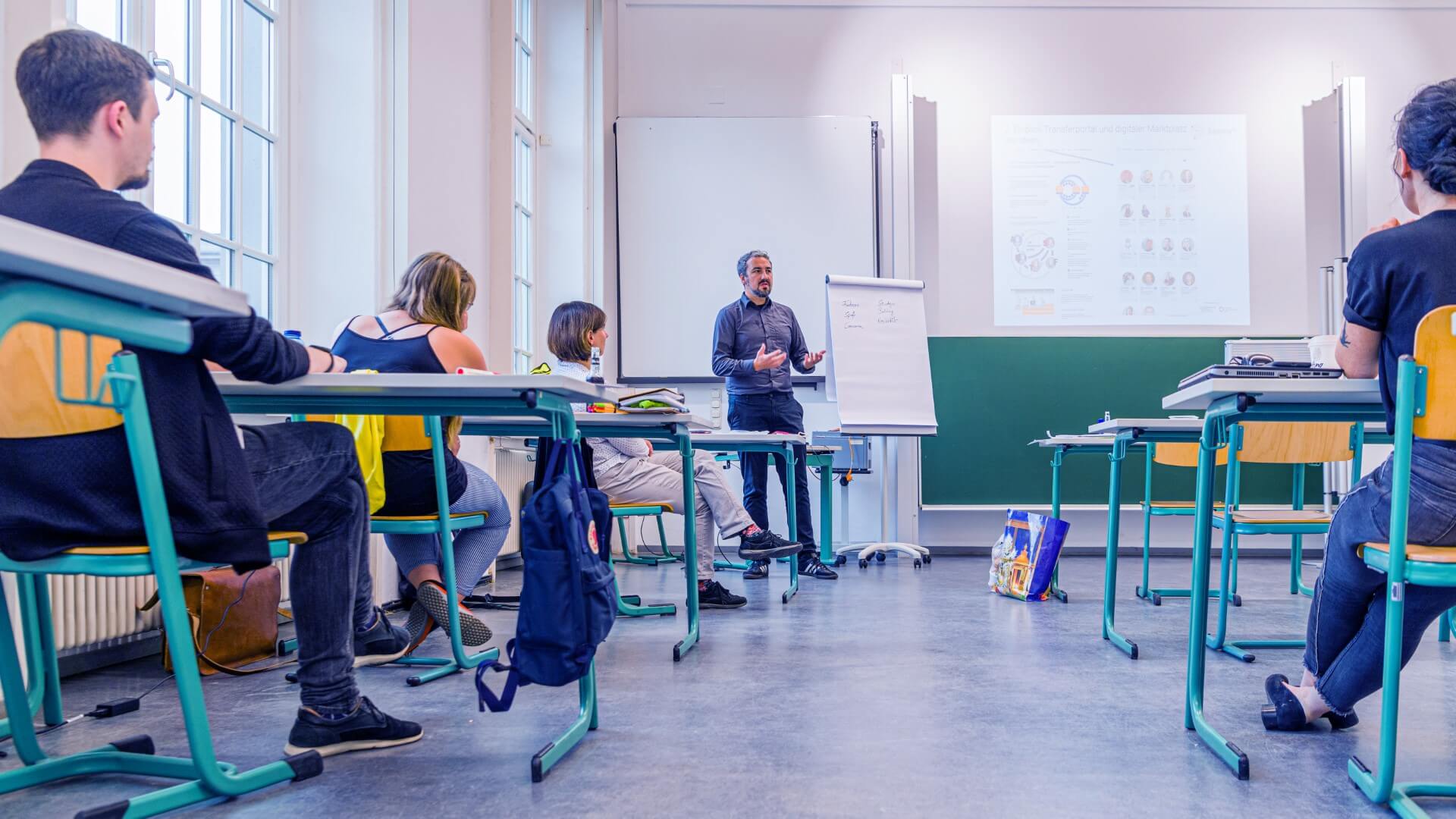 Stehende Person erklärt sitzenden Personen etwas in einem Unterrichtsraum.
