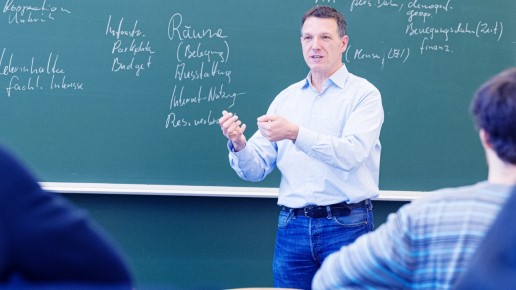 Männliche Person steht vor einer Schultafel auf der Begriffe stehen.
